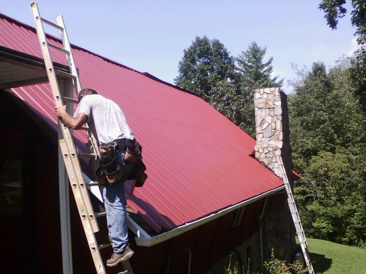 Roofer on a ladder beside a red metal roof and stone chimney — Soffit & Fascia Repair by Roofing Brothers in Copeland